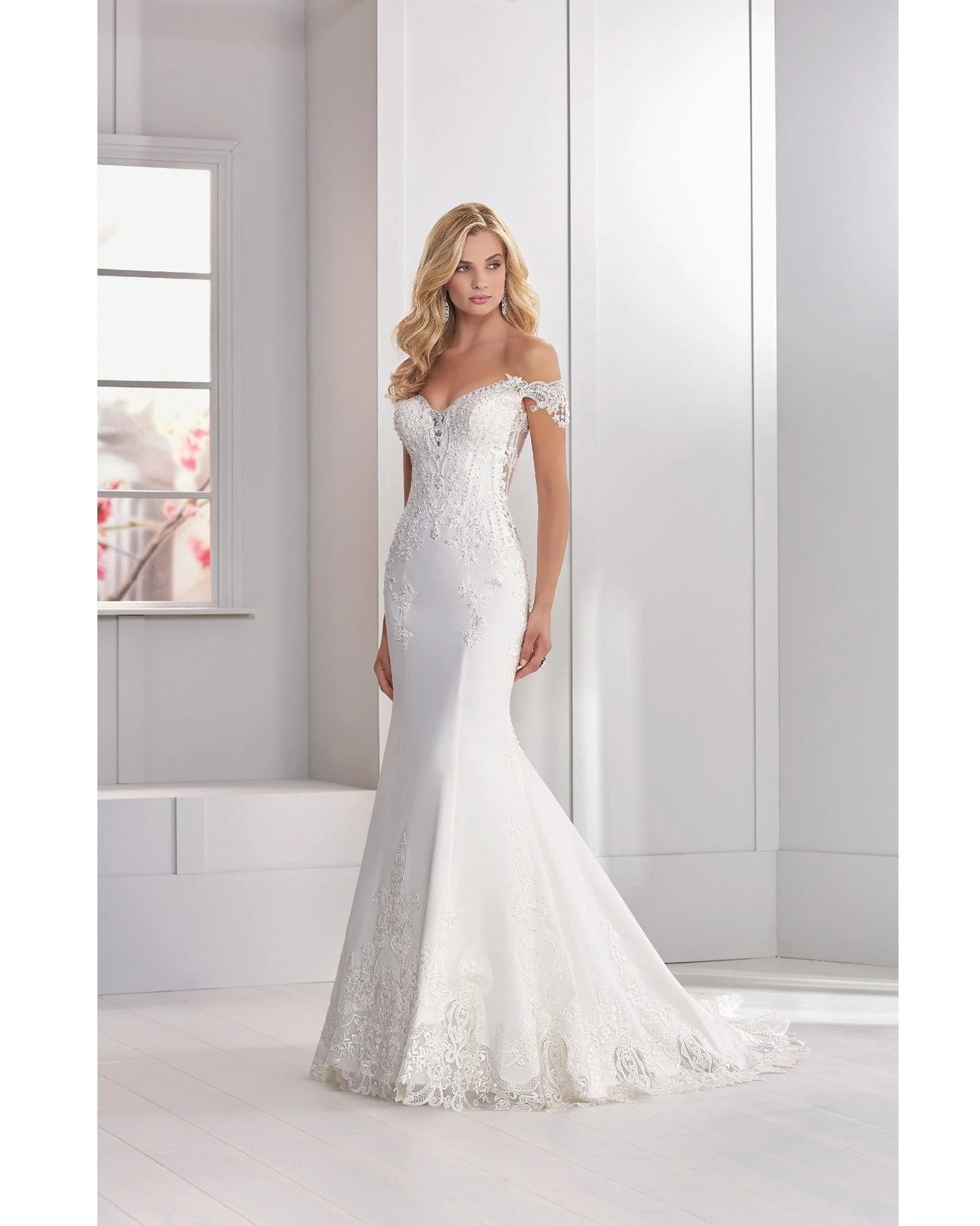 A woman in a white wedding dress with lace details and a fitted silhouette, standing in a bright room.